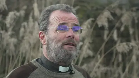 Paul Cawthorne, who is wearing glasses, a jumper and clerical collar, is standing in a field as he is being interviewed. 