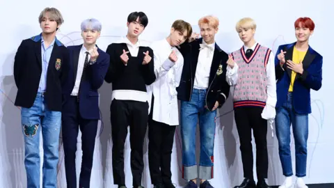 Getty Images From left to right, V wears blue jeans with a butterfly on the thigh and a dark suit jacket, he has light silvery hair. Suga wears a navy suit with a tie, he has light purple hair. Jin wears black trousers and a black sweater with white shirt, he has black hair. Jung Kook wears a long white shirt and black trousers, he has brown hair. RM has a white shirt and black suit jacket with jeans, he has light orange hair. Jimin wears a pink, red and navy sweater vest with white shirt and black trousers, he has blond hair. J-Hope has jeans and a yellow t-shirt with navy jacket on, he has ginger hair. 
