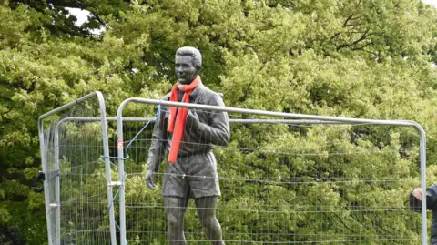 Fenced off statue of Brian Clough wearing a red scarf. There are trees in the background.