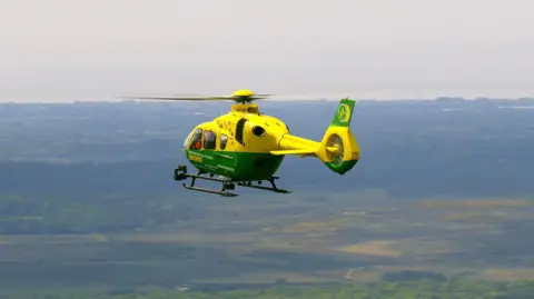 A green and yellow ambulance helicopter is flying mid-air against a backdrop of green land and the coast.