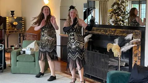 Lauren Hutchinson and Rachel Stammers are both wearing 1920s fringed flapper dresses and are practicing the Charleston in front of a large fireplace.