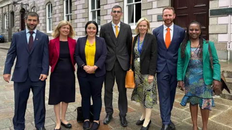 BBC Seven people are standing together outside a large stone-fronted building with tall windows and a dark wooden doorway, posing for a group photograph on a paved street.
The group is dressed in formal business clothing, including suits, jackets and dresses, with several people wearing brightly coloured jackets or ties, suggesting the photo may have been taken at an official or professional event.