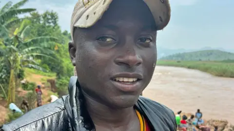 Congolese man Jordan Bita wearing a beige cap and a wet black waistcoat. He is standing on the Burundi side of the River Rusizi - its brown roaring waters can be seen behind him.