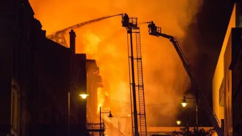 Getty Images Two firefighters on ladders douse a large fire with water. It is dark and the flames and smoke have created a large orange glow in the background of the image.