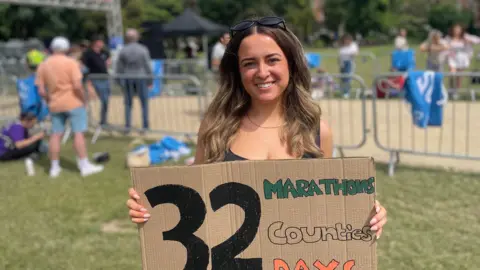 A smiling woman stands outdoors at what appears to be a running event, holding a large cardboard sign that reads: “32 MARATHONS Counties DAYS” in hand-painted black, green, and orange lettering. She has sunglasses resting on her head, and behind her are metal barriers, scattered spectators, trees, and an event banner under a cloudy sky.