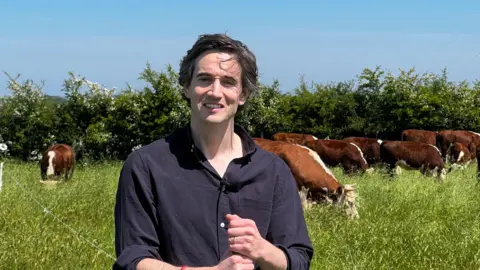 Farmer Aidan Stanley stands in a field near Bath while behind him a small herd of brown and white cows is visible. The grass in the field is very tall, and there is also a hedge in the background