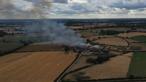 an aerial image of an agricultural farm in the middle of many fields is on fire. the flames are large and orange with grey smoke billowing from them and into the air