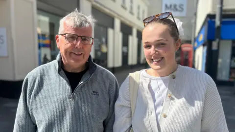 Brian and Freya Johnson stand next to each other by some shops. Brian wears a grey zip-up jumper, has short grey hair and wears black spectacles. Freya wears a white cardigan and brown sunglasses on her head. Her brown hair is tied back with a large white scrunchie.