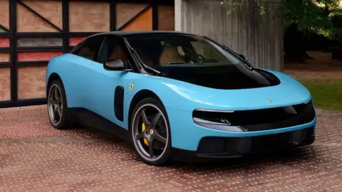 A light blue electric Ferrari parks in front of a garage