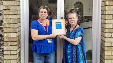 Salvation Army Trading Company Two women, both dressed in blue are standing in front of a shop and they are holding up a book for the camera.