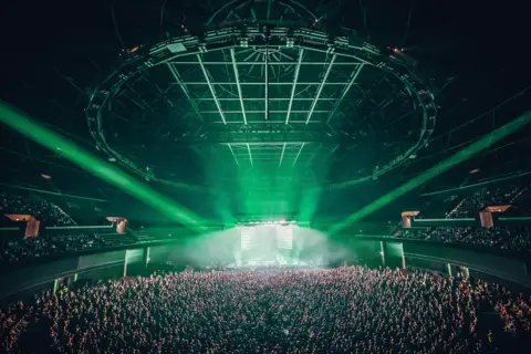 Hydro Inside shot of the area showing its vast space. Thousands of fan stand in the pit and in the seating to the sides. Green lights shine on the stage. 