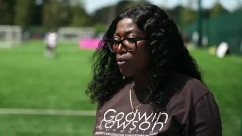 A lady in a brown T-shirt and sunglasses with long dark wavy hair is standing on a football pitch being interviewed for TV.