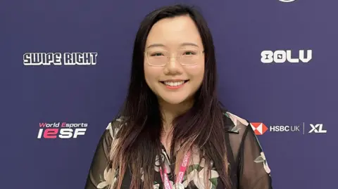 Dr Lucy Zhu A woman is smiling in front of a branded background with "World Esports IESF" written on it alongside sponsors