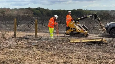 National Highways Two men wearing yellow and orange high-viz are working net to a fence. One man is operating a mechanical drill. The land around them has been burnt.