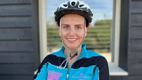 A young girl wearing a bike helmet and a blue and black jacket smiles into the camera.
