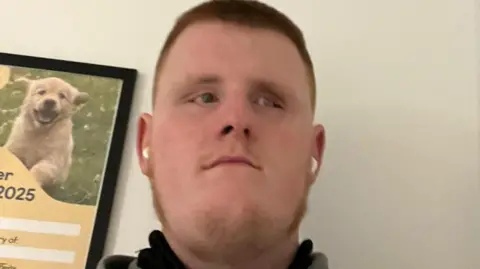 Tiernán Devine Tiernán Devine has short brownish red hair and has stubble. He is wearing white airpods and is in a grey sweatshirt. He is standing near a white wall that has a Guide Dog Poster in the background with a picture of a very happy looking golden retriever.