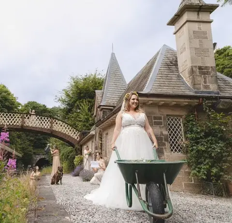 Lucy Heath A woman in a wedding dress pushing a wheelbarrow at a heritage rail station