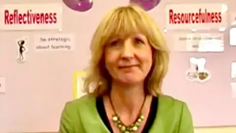 Family Christine Baker is seen in front of a school noticeboard showing a display about learning strategies. She has blonde hair with a fringe and is wearing a green jacket with a black top and a necklace of green beads.
