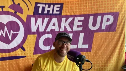 Joe Sims is wearing a yellow t-shirt, glasses and baseball cap. He is sat behind a radio microphone in front of a banner that says 'The Wake Up Call'.