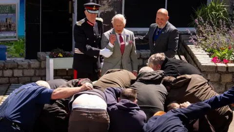 PA Media King Charles III is held back by a man in uniform and a man with a bald head as a group of men grapple for a ball in front of him