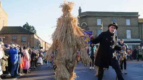 PA Media The Straw Bear is paraded through the streets accompanied by attendant keepers, musicians and dancers during the Whittlesea Straw Bear Festival in Whittlesea, Cambridgeshire on 13 January 13 2024. Leepers is leading the Straw Bear by some rope and wears a full black uniform and hat. 