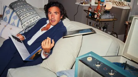 Jack Savoretti sitting on a white couch with white and colourful cushions. He is holding a vinyl record and is looking with surprise at the camera. There is a cart with alcohol behind him.