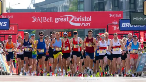 A row of men wearing running bibs and shorts setting off on a race, with people either side watching them. A large red banner with the words "AJ Bell Great Bristol Run" is visible behind them