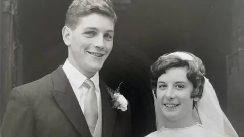 A black and white photo of Anthony and Helen Chandler at their wedding in 1966. Anthony is on the left and has short hair and is wearing a shirt, tie and suit jacket. Helen is on the right and is wearing a white veil and dress.