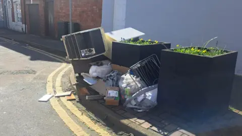 A pile of dumped household waste sitting on a street corner beside two large planters with yellow flowers. The rubbish includes a broken appliance, cardboard, bags of clothing and metal frames. The contrast between the overflowing fly‑tipped items and the neatly maintained planters highlights the impact of illegal dumping in an otherwise tidy public space.