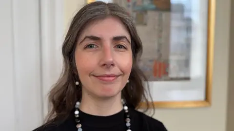 Ashleigh Hibbins, a woman with long wavy brown hair, standing in front of a framed picture. She is wearing a black top and a necklace.