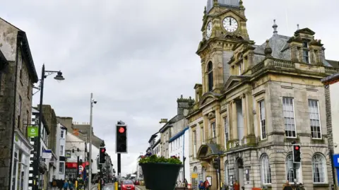 Kendal Town Hall. It is an ornate stone building with a clock tower. Shops and traffic lights can be seen on the opposite side of the street.