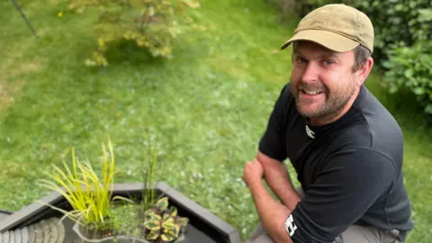 Michael Topping wears green shorts and a black top. He has a green cap. He is kneeling beside a water feature. There is grass and shrubs around him.