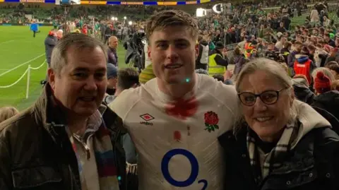 From left-to-right, Cliff Freeman with short grey hair wearing a brown jacket, Tommy Freeman with short dark hair wearing a white England shirt with an O2 logo on the front, and Sara Freeman with short light-coloured hair and glasses wearing a dark jacket with white hood. Tommy has his arms around them and they are standing in a sports stadium with a crowd of people behind them. The rugby pitch is visible to the left.