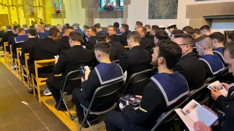 Sailors n blue uniforms sit in rows inside the church holding copies of an order of service