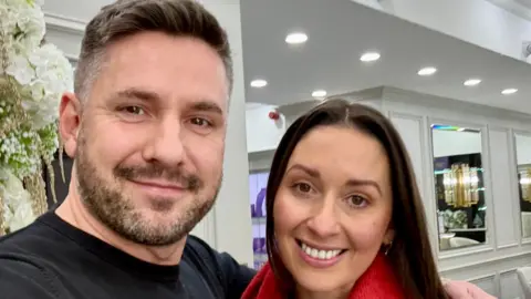 David Challinor A man and woman pose indoors for a selfie, smiling. The man has a short and neat haircut, stubble and wears a black t shirt. The woman has dark brown hair and eyes and wears a red scarf.