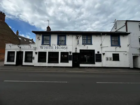 A large, white tow-storey pub beside a two-lane road. Ther are cloudy skies above it. The pub has 12 large windows and two doors. The pub's name, "The White Horse" is written on the front.