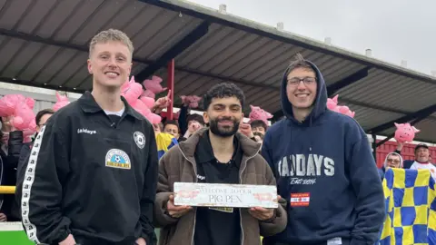 Connor Bennett/BBC Ellis Platten (left) and Alfie Indra (centre) with Ben Gibbs on the pitch at Witham Town. Alfie is holding a sign up in front of him saying welcome to our pig pen. Behind are the stands with men carrying pink pigs, yellow and blue flags and cheering. 