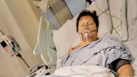 Giuliana Rossi Guiliana is sleeping in a hospital bed. She is wearing a blue nightgown and a breathing mask over her nose and mouth. She is sleeping and is connected to several tubes.