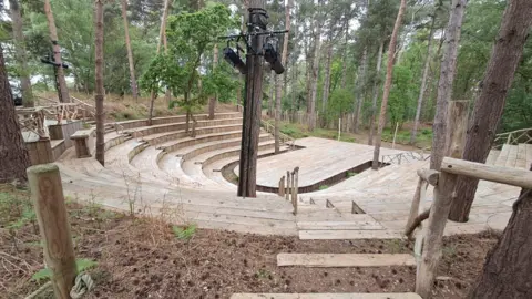 Darren Rozier/BBC A view of an outdoor theatre with raised seating on incremental levels. The stage and seats are made of wood and are surrounded by trees. 