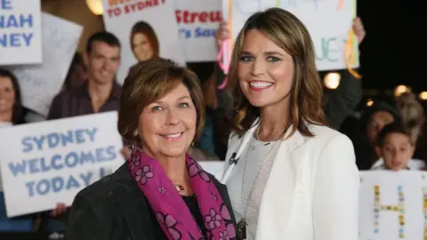 Nancy Guthrie is on the left and Savannah Guthrie on the right, on the set of NBC's morning show Today. There are people in the background with 'Welcome to Sydney' banners. Nancy is smiling and wearing a purple scarf while her daughter is also smiling, wearing a white suit