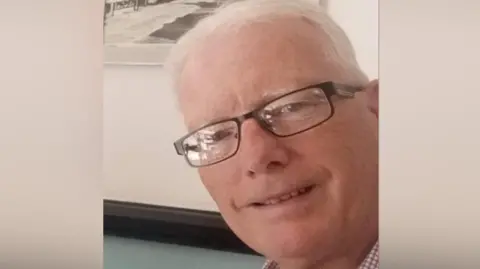 Andrew Avery smiling for the camera. He is wearing glasses. There is a picture on the wall behind him.