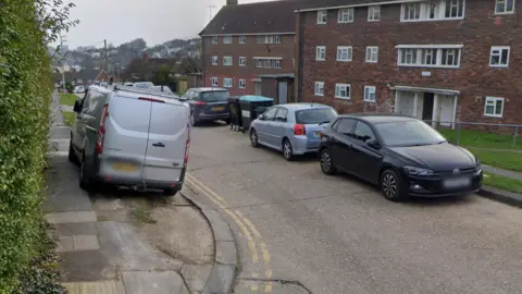 A silver van is parked with four wheels on a pavement marked by double yellow lines at the junction of Tintern Close and Horton Road. Cars line the other side of the road, with no yellow lines, leaving limited space for cars to pass through.