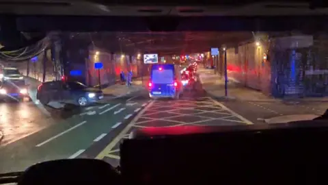 Rob Cartwright A dark blue van with its break lights under a railway bridge stuck in traffic. A green cycle lane can be seen to the left and a lit pathway on the right. The picture has been taken from the cab of a fire engine.