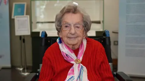 Joan Ramsey - a lady sat in a red knit jumper with an elegant scarf. She is wearing glasses and looking directly at the camera