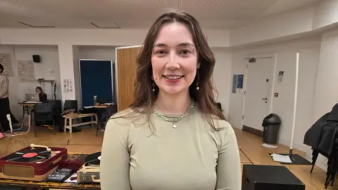 Tamzin is wearing a khaki long-sleeved top and has long brown hair. She is also wearing two necklaces and is smiling at the camera as she stands in a room with other students who are taking part in a lesson behind her.