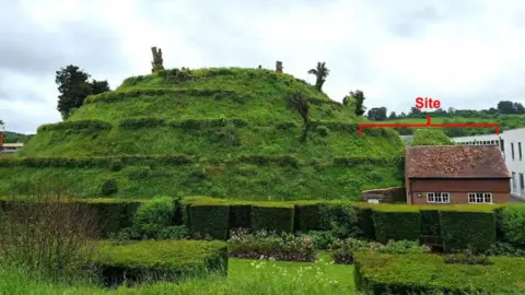 Marlborough College/Marlborough Mound Trust A side view of the Marlborough Mound. It is a grassy hill with six rings cut into its side. It has a few trees on top and is surrounded by buildings - one of which is in clear view to its right.