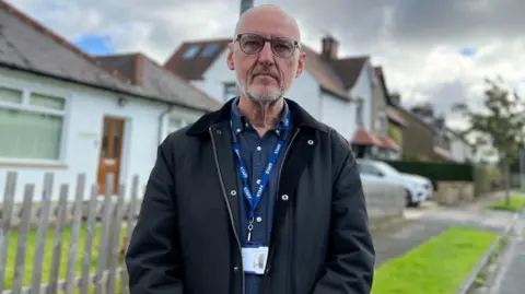 Aisha Iqbal/BBC A man stands in a pretty looking suburban street with white painted houses and neatly trimmed grass verges visible  in the background. He is middle aged, has a shaven head, a short, stubbly white beard and black glasses. He wears a dark blue shirt, and dark zip up jacket with collar.