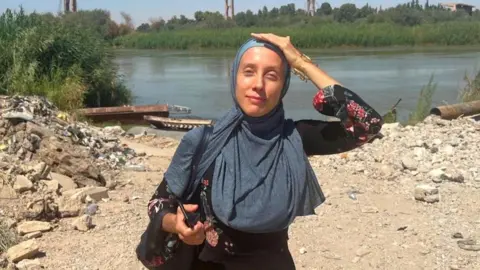 An undated image of Shelly Kittleson. She wears a hijab and holds a phone while standing in front of a river