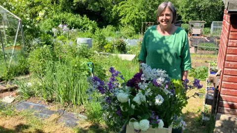 A woman with mid-length greyish hair and wearing a green top stands in what looks like an allotment, with lots of plants and trees around. There is also a greenhouse and a shed. She is pushing a wheelbarrow which contains brightly-coloured flowers.