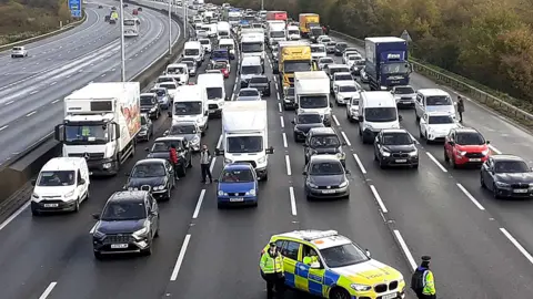Just Stop Oil/PA Six lanes of held traffic on a motorway. There is a police car in the foreground and lots of vehicles behind the police car.